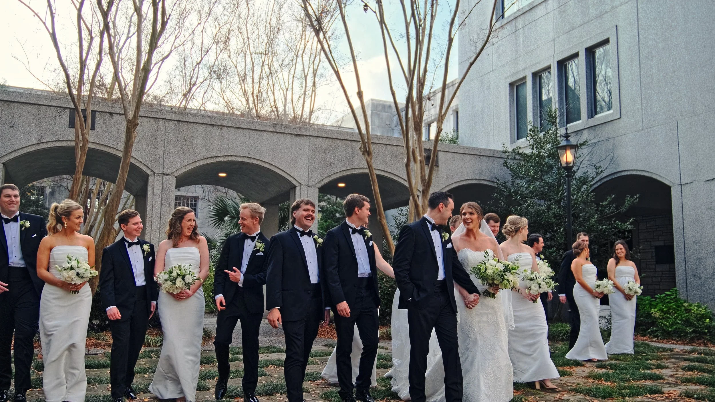 Group of wedding party members walking outdoors, dressed in formal wedding attire, with bridesmaids in white dresses and groomsmen in black tuxedos, holding bouquets and smiling.
