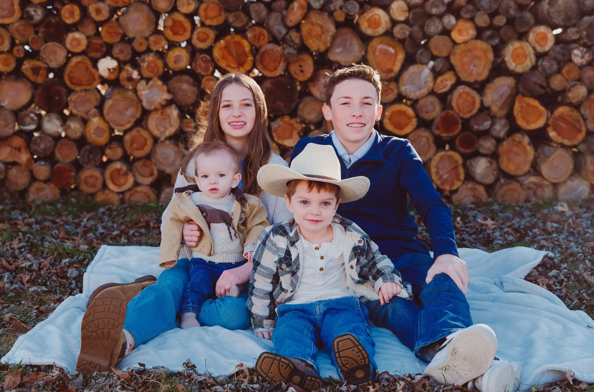 A family of five sitting on a white blanket on the ground in front of a large stack of chopped logs, outdoors, during daytime. The group includes a teenage girl, two young boys, a young girl, and an older boy, all dressed in casual outdoor clothing.