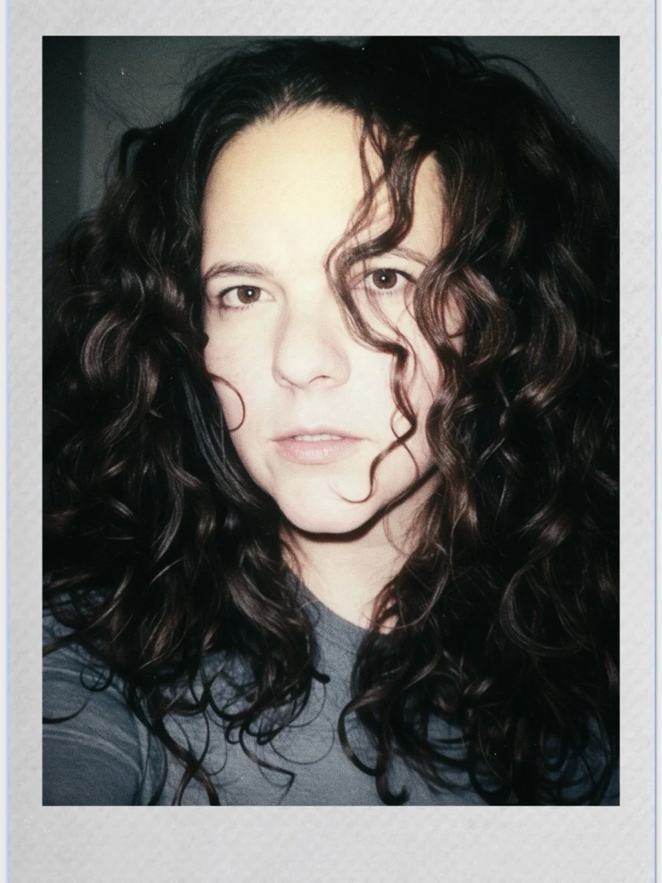 Close-up portrait of a person with long, dark, curly hair and light skin, looking directly at the camera.