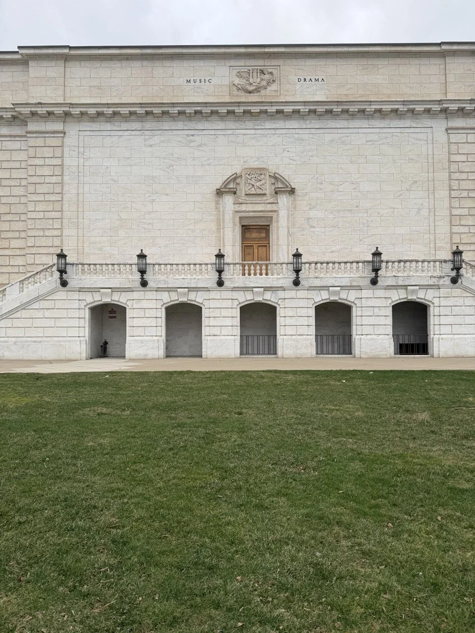 The exterior of a building with a stone facade, decorative architectural elements, black lamps, and a central wooden door. A grassy lawn is in the foreground.