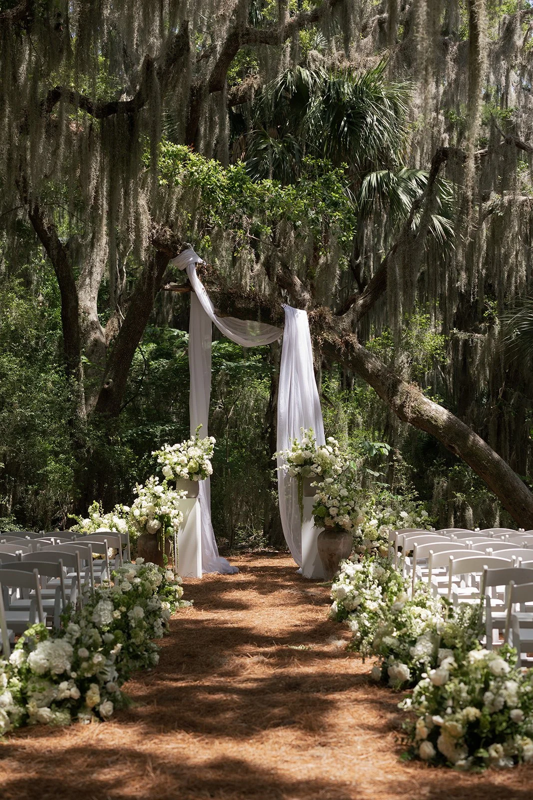 outdoor walkers landing wedding aisle with green and white flowers, white chairs, and a floral arch draped with white fabric in a wooded area with Spanish moss hanging from trees. large white and green urn arrangements framing the alter