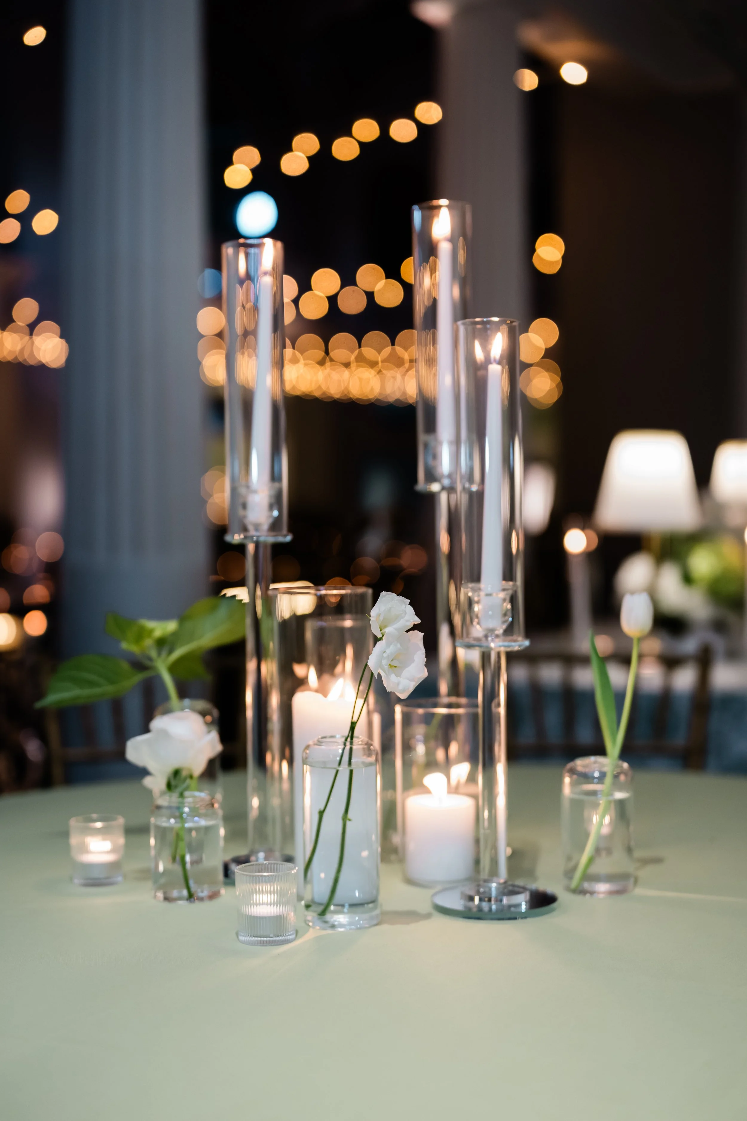 A centerpiece of  raised taper candles and bud vases on a table at a lightner museum wedding , with string lights in the background.
