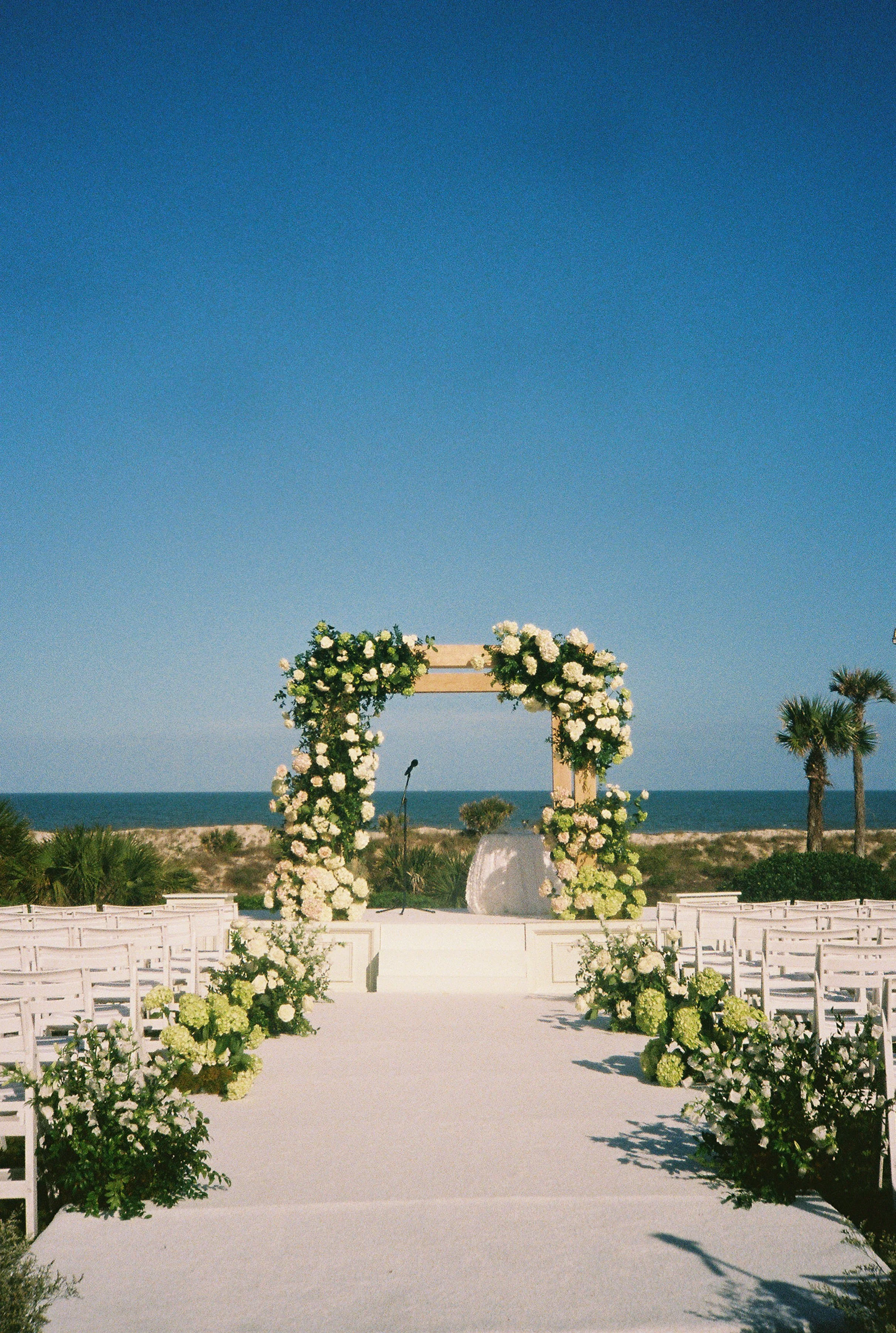 A wedding ceremony setup on the lawn at the ritz carlton, with a fully globalized chuppah, and white chairs, overlooking the ocean and palm trees under a clear blue sky. clustered arrangements line the aisle like a meadow