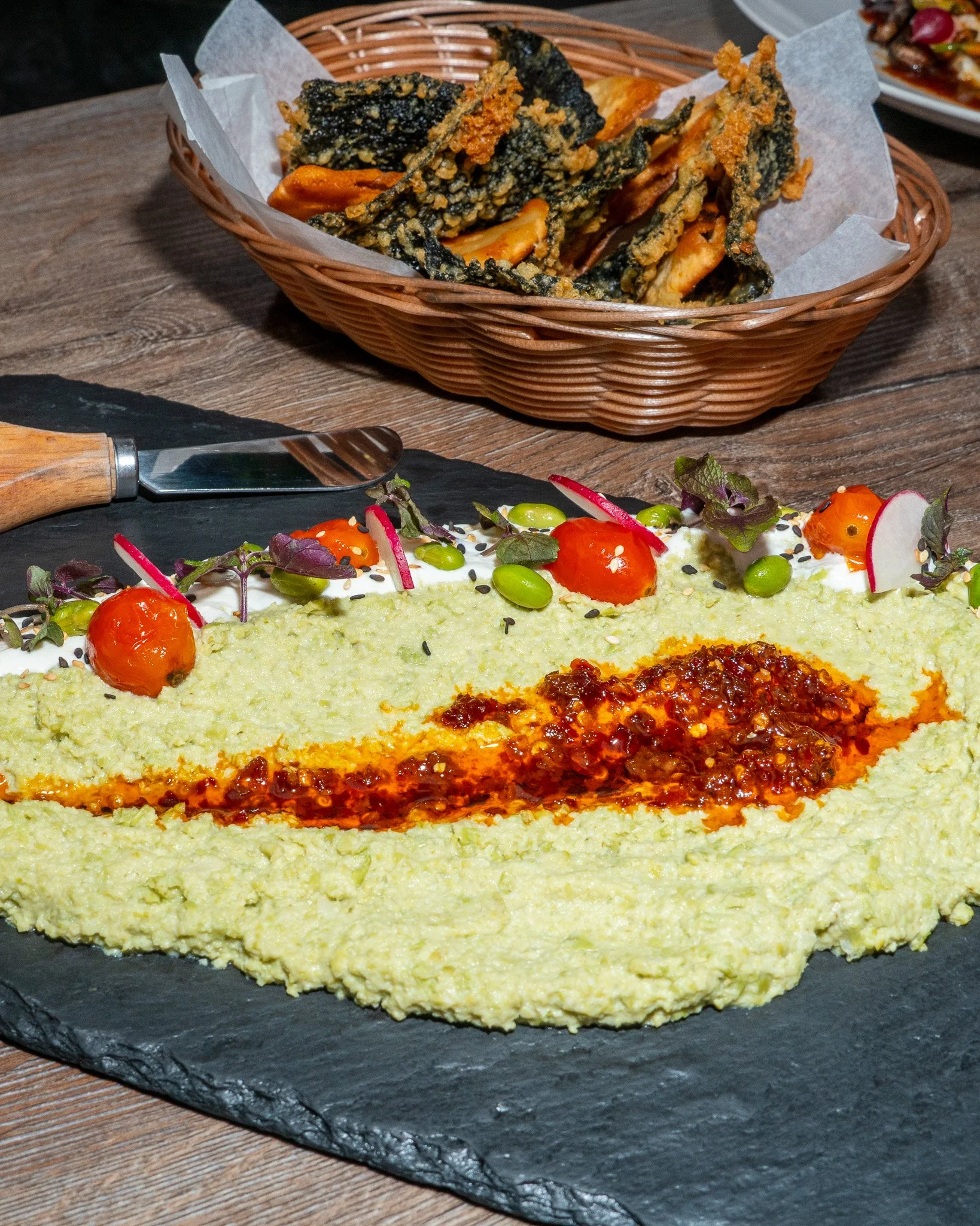 A black slate platter with a large serving of hummus garnished with cherry tomatoes, radishes, edamame, and herbs. In the background, there is a basket of battered fried eggplant slices on a wooden table.