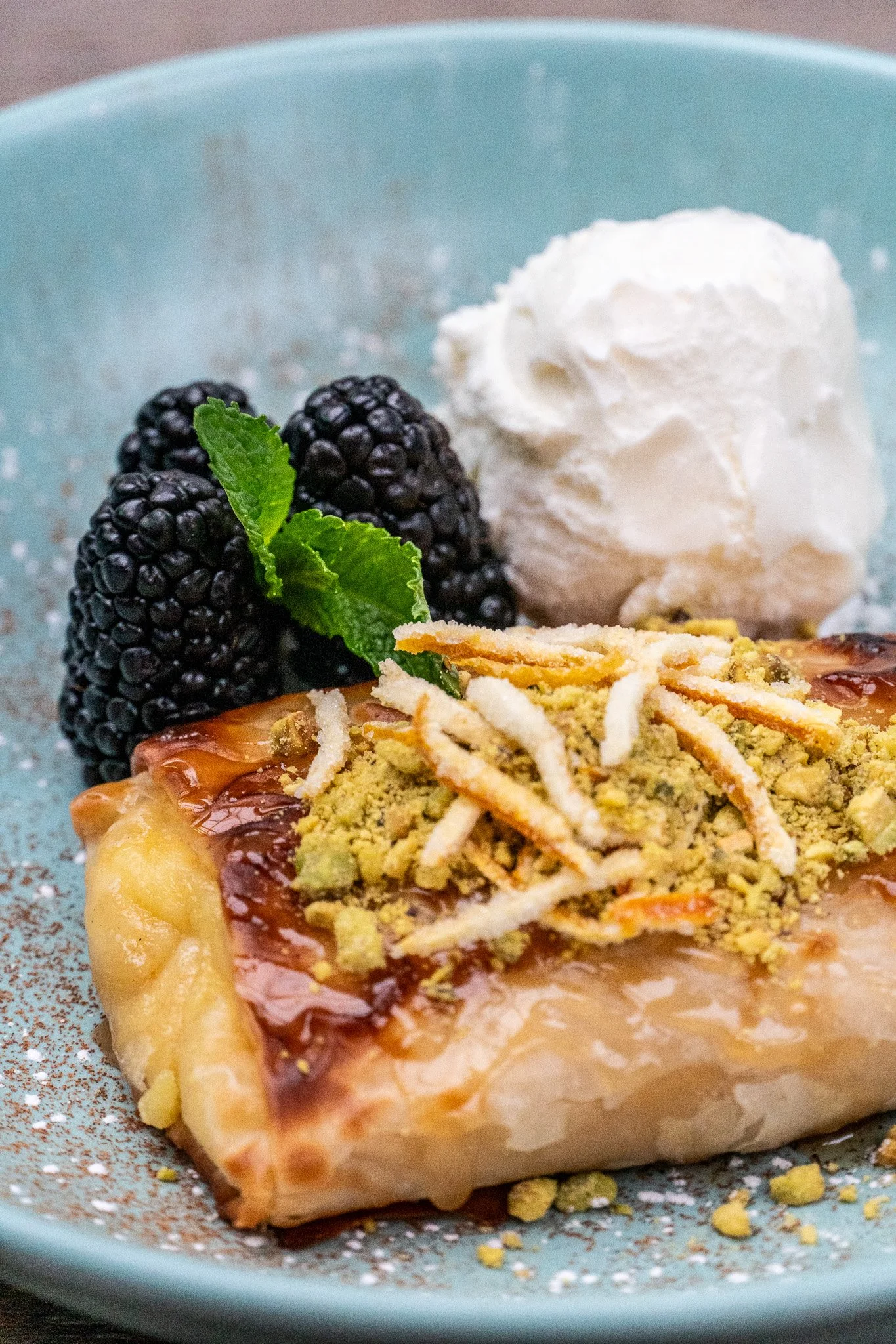 A slice of dessert topped with crushed nuts and white strips, served with blackberries, mint leaves, and a dollop of whipped cream on a blue plate.