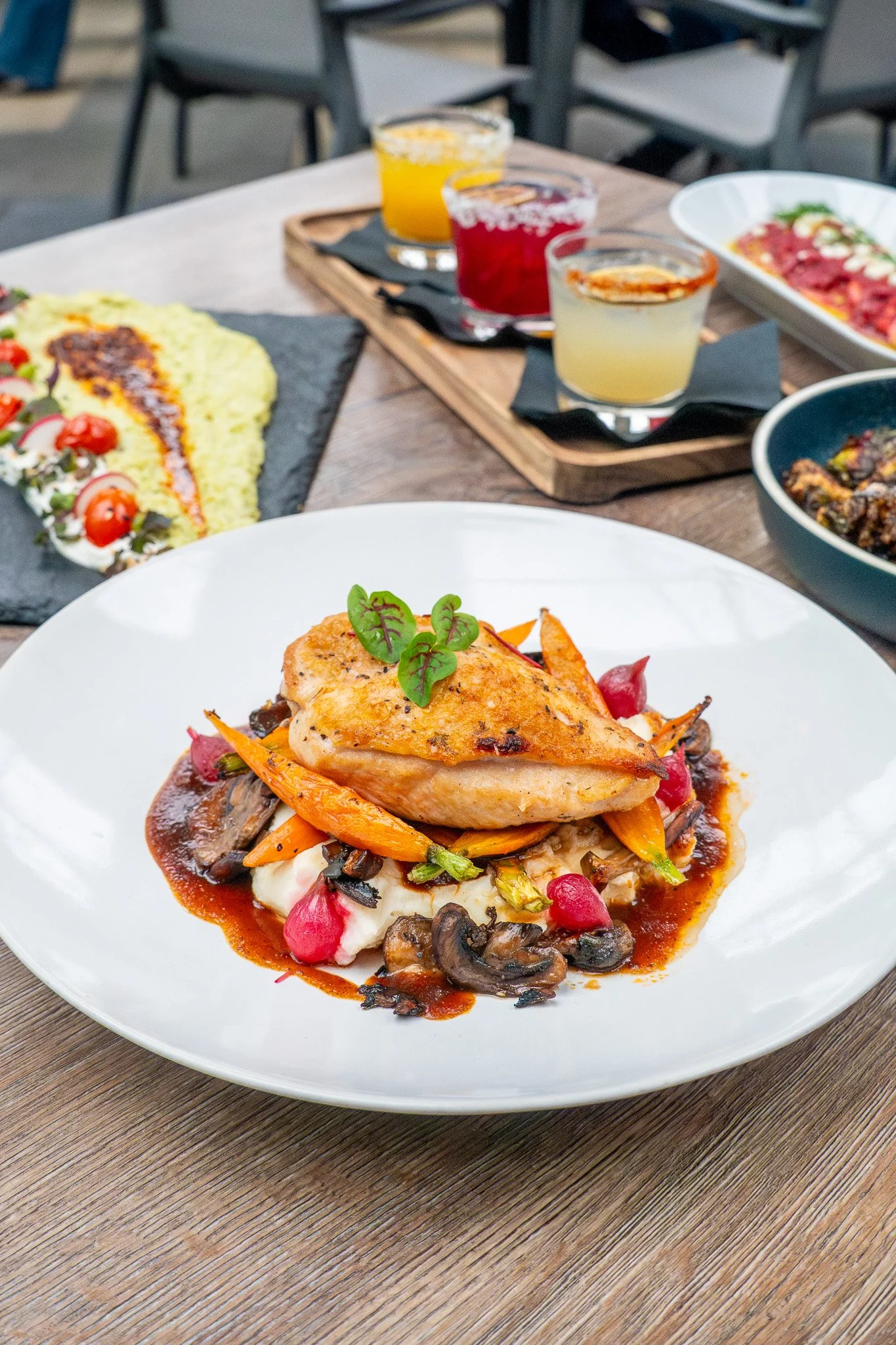 A plated gourmet chicken dish with vegetables and sauce on a white plate, with a variety of colorful beverages in the background on a wooden table.