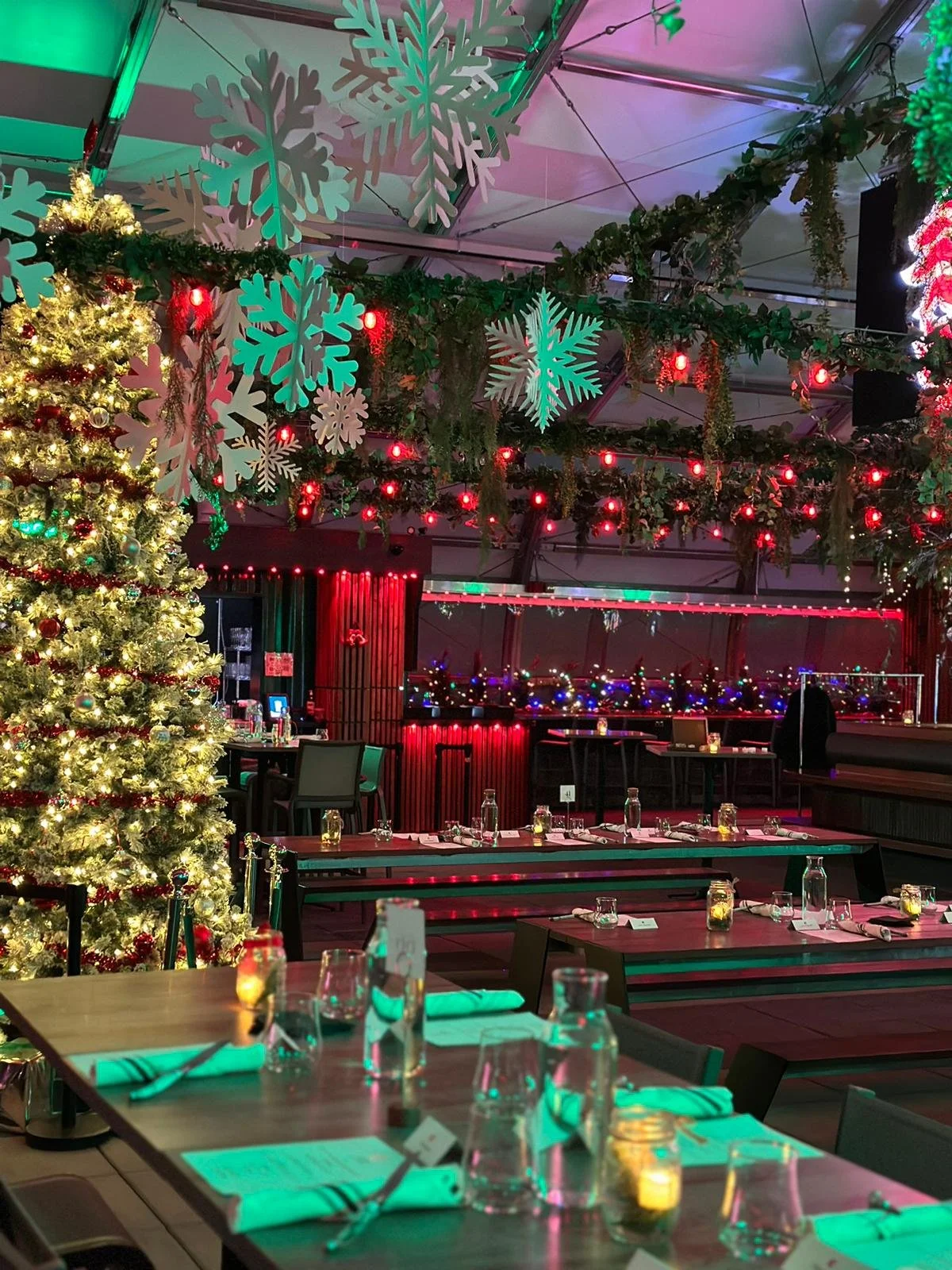 Indoor space decorated for Christmas with Christmas tree, large paper snowflakes, and colorful string lights, set up for dining or celebration.