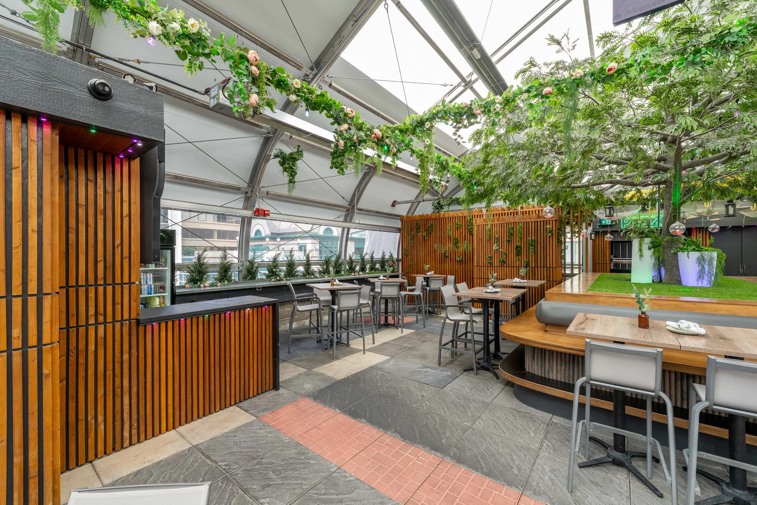 Indoor restaurant with outdoor garden theme, featuring large trees, potted plants, and hanging greenery, with tables and chairs arranged for dining under a curved glass ceiling.