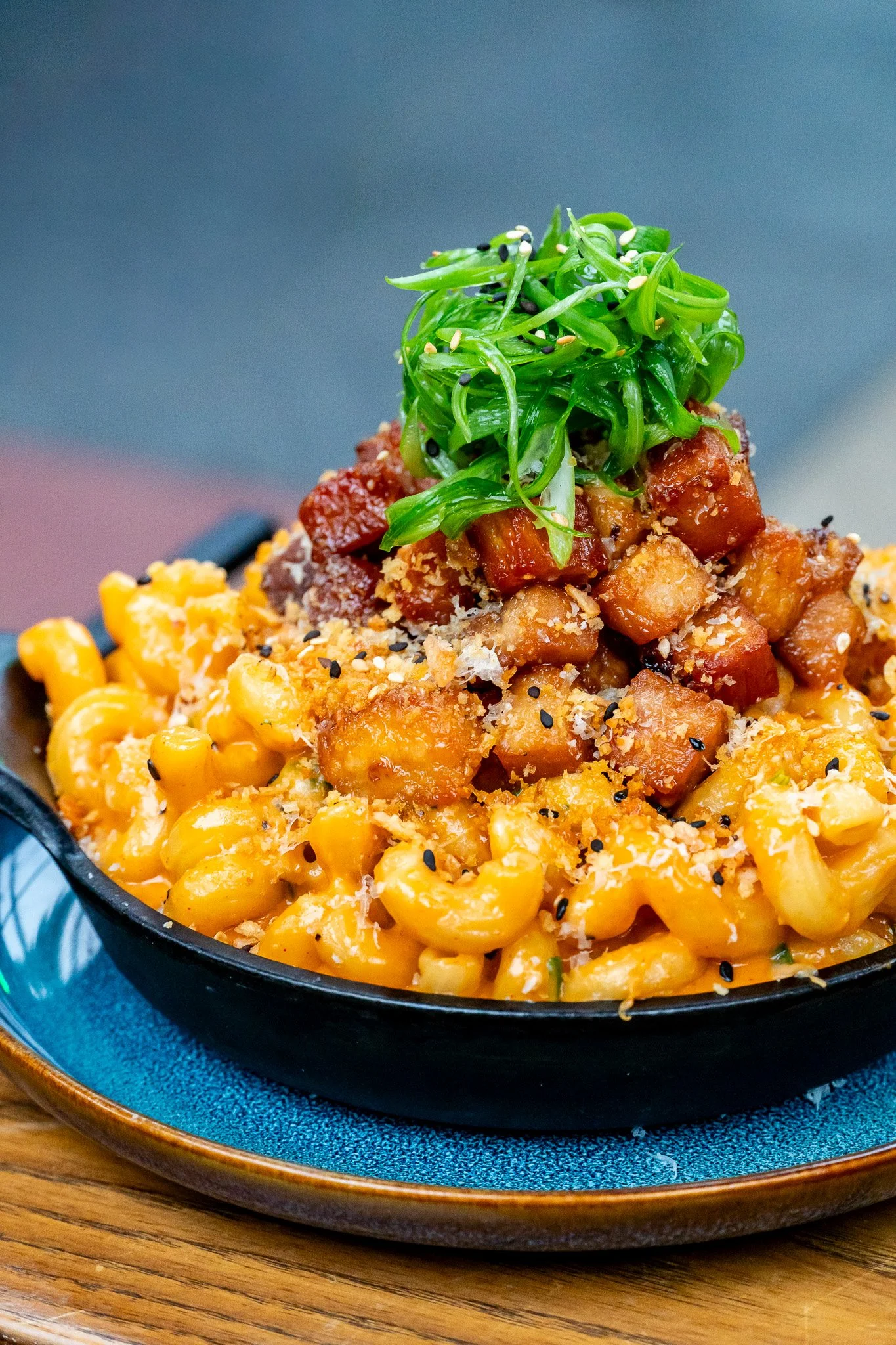 A black skillet filled with macaroni and cheese topped with chopped pork belly, green seaweed salad, sesame seeds, and grated cheese.