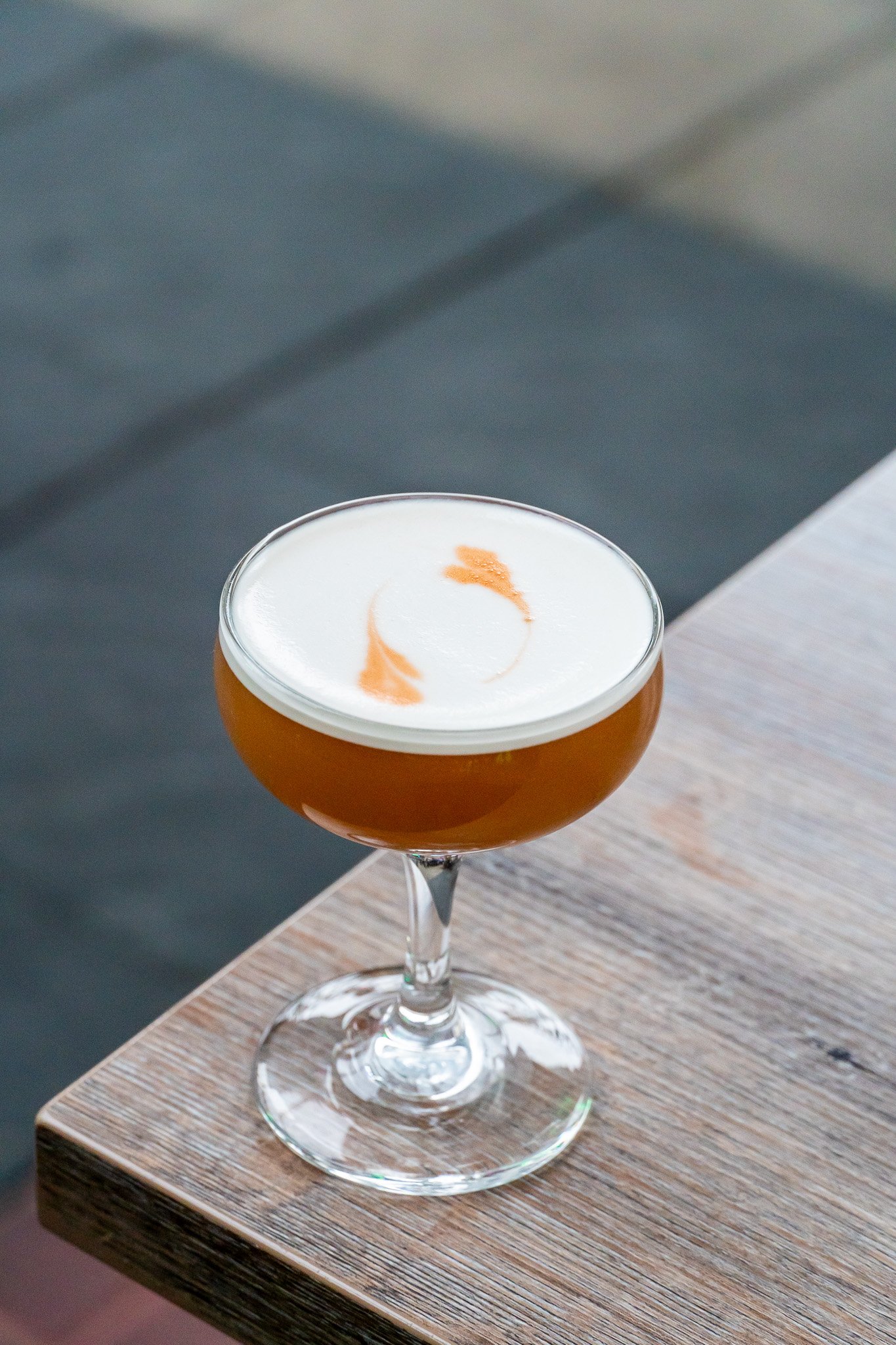 A cocktail in a coupe glass with a light brown liquid and a foamy white top, garnished with an orange peel curl, placed on a wooden surface.