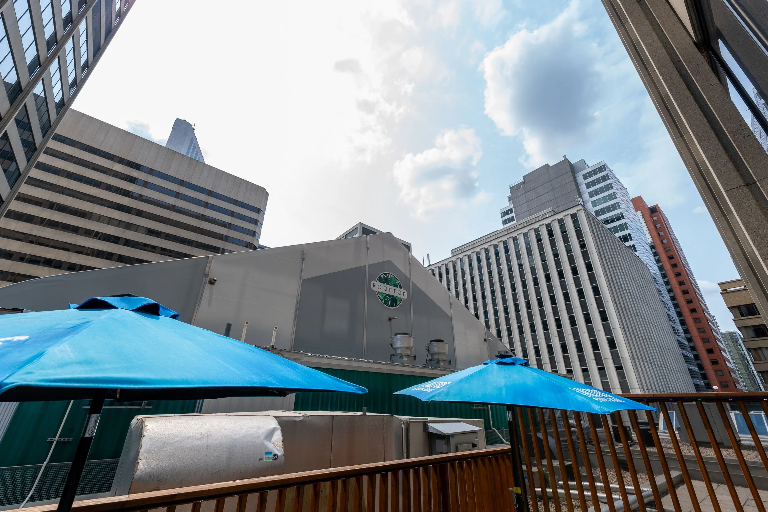 Cityscape with tall buildings, blue sky, and clouds, seen from a rooftop area with blue umbrellas and a wooden railing.