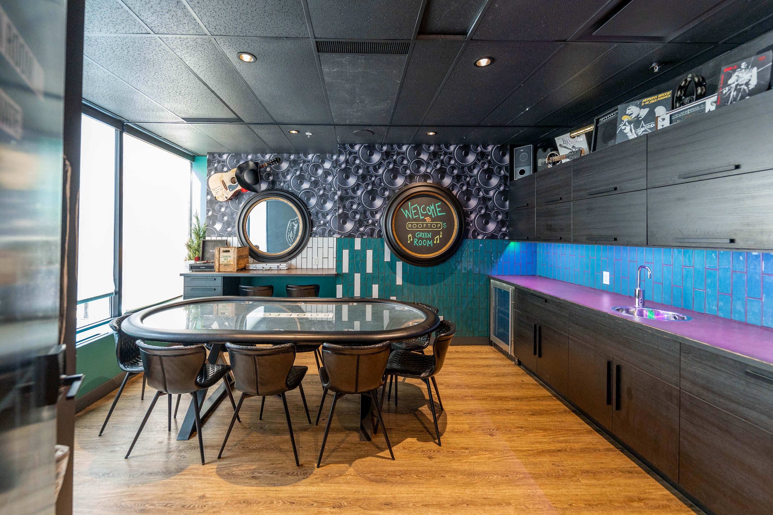 Interior view of a modern rooftop lounge with a long purple countertop, a sink, black cabinets, a glass-top card table with black chairs, decorative wall with circular patterns, a chalkboard sign that says 'Welcome to Rooftop Green Room', guitars, and large windows.