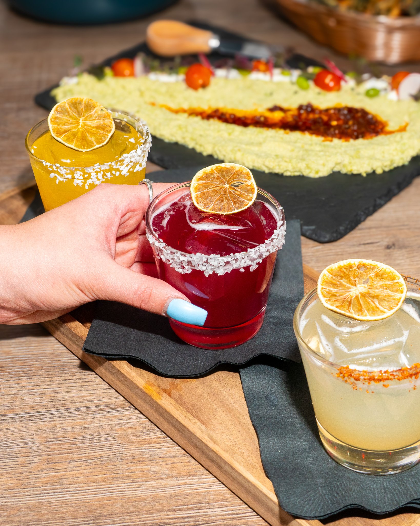 Three colorful cocktails topped with dried orange slices on a wooden table, with a Mexican dish called 'torta de tamal' in the background.