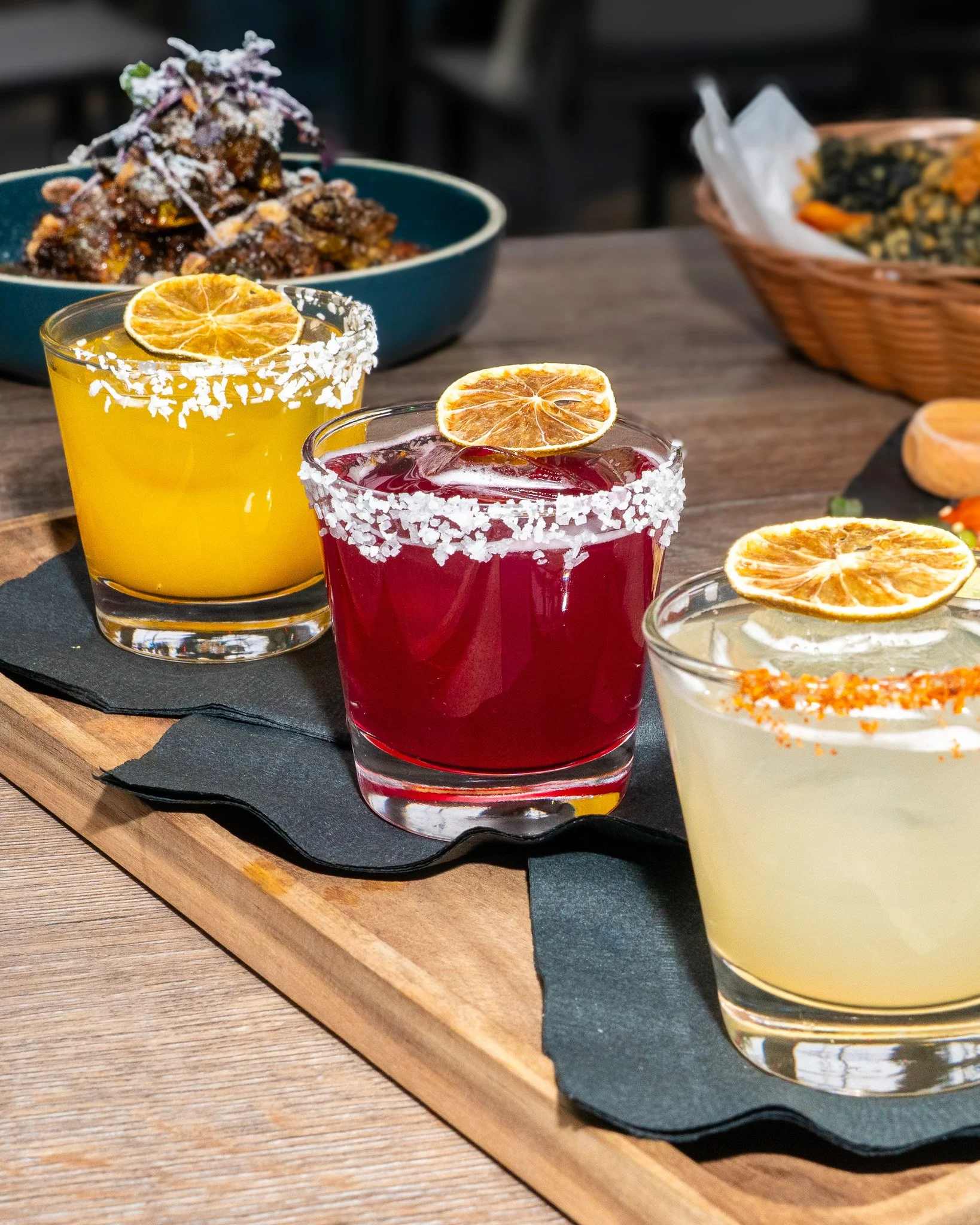 Three colorful cocktails garnished with dried orange slices on a black napkin, on a wooden serving tray.