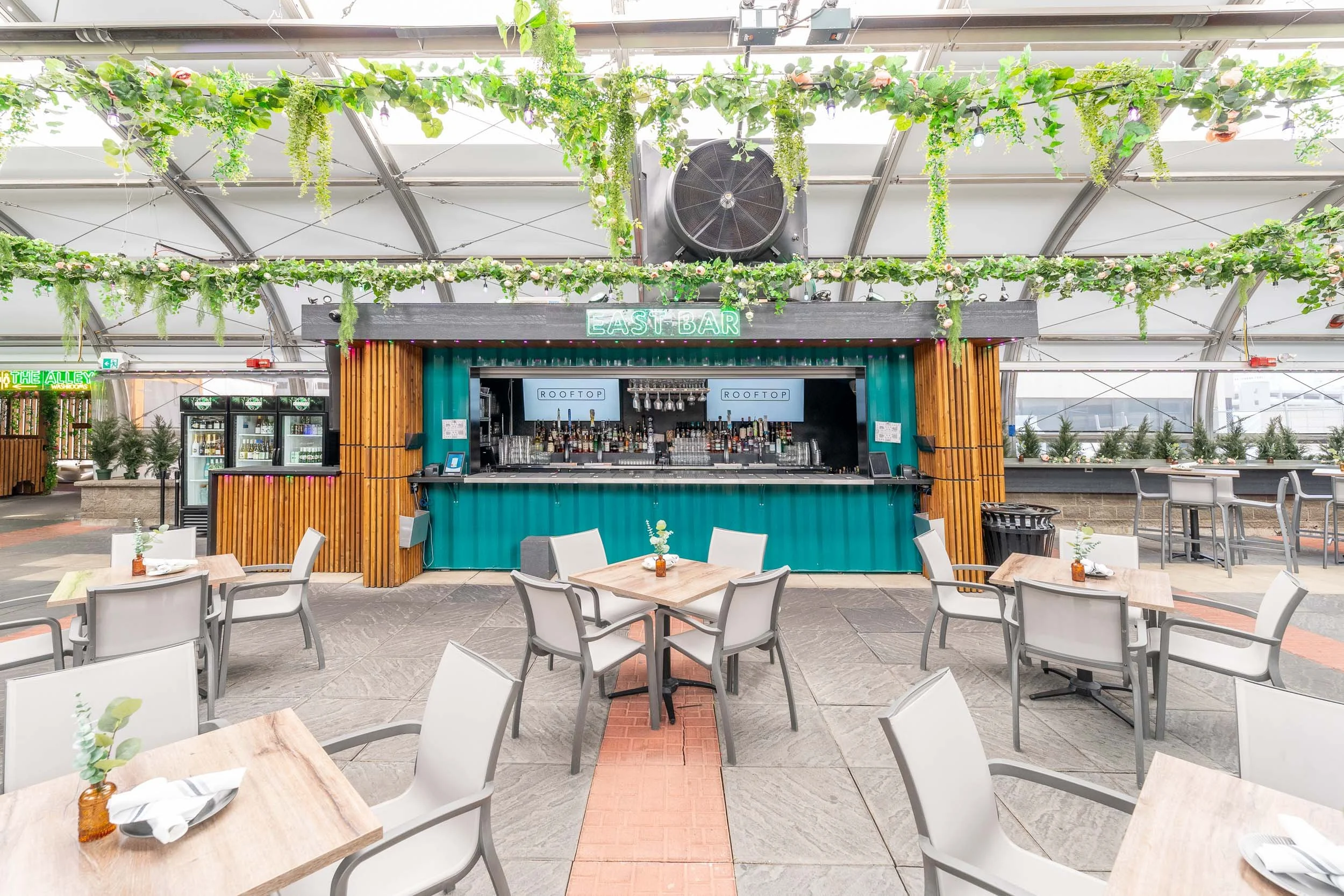 Indoor rooftop bar with a central serving area, surrounded by tables and chairs, decorated with greenery hanging from the ceiling, and a neon sign reading 'East Bar' above the bar.