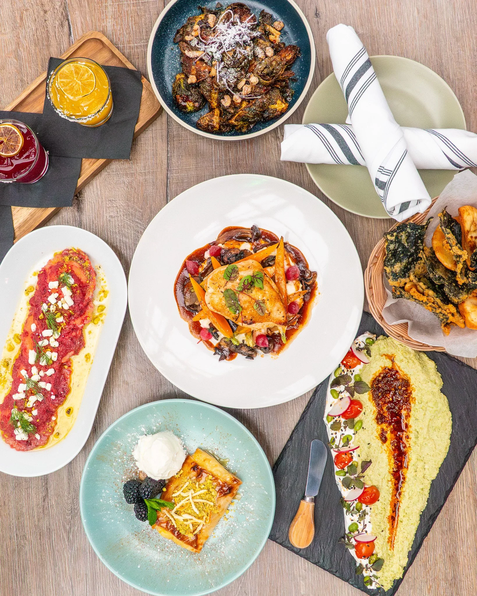 A variety of gourmet dishes including a colorful rectangular cake with berries and cream, a white plate of meat with vegetables, a black slate serving of mashed potatoes with vegetables, a white oval plate with a colorful tomato and fish dish, a bowl