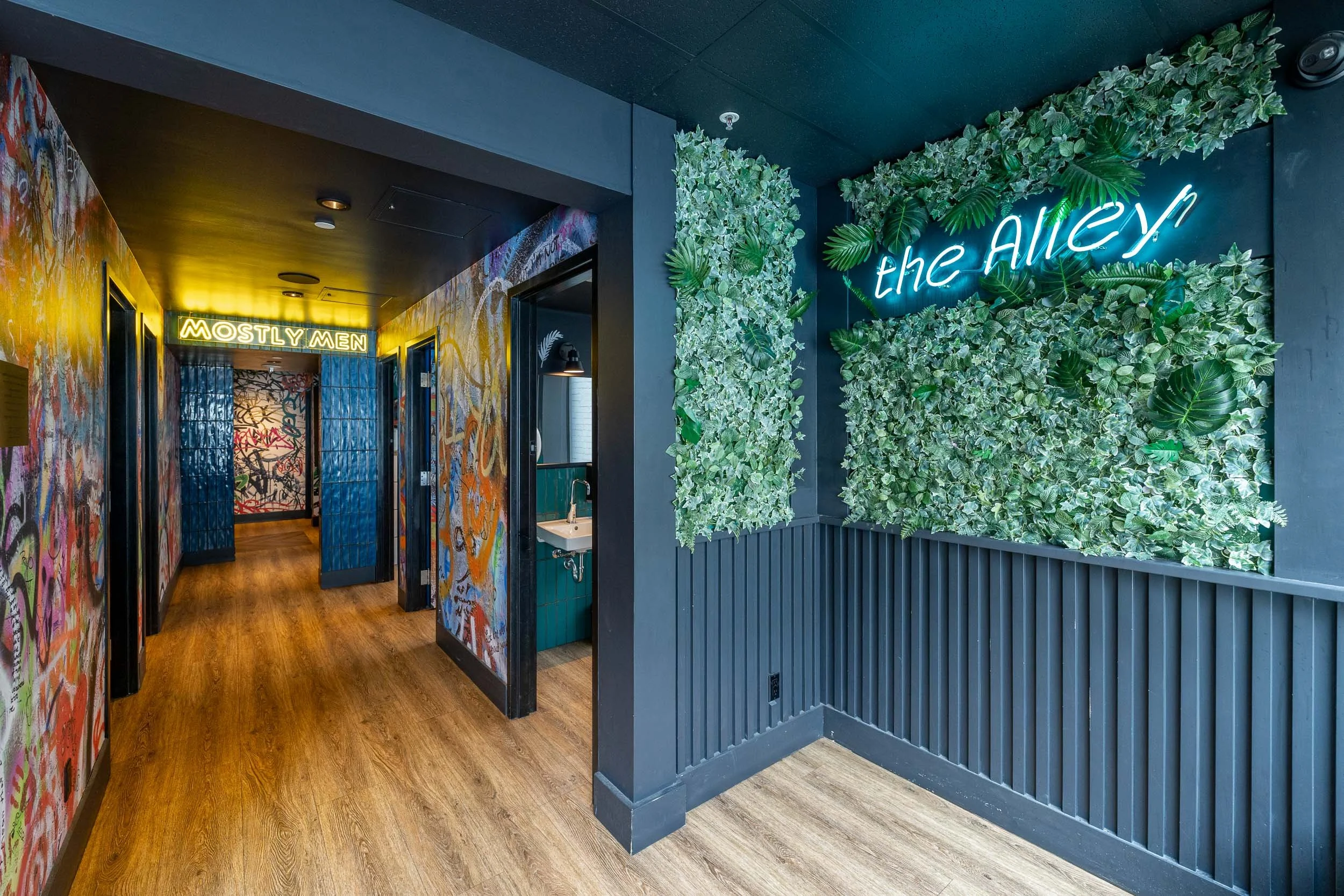 Interior of a modern restroom with graffiti-covered walls, wooden floors, and a green leafy wall with neon signs reading "the ALLEY" and "MOSTLY MEN."