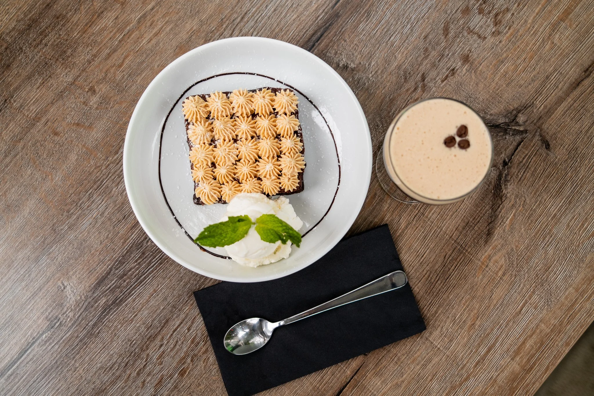A dessert plate with a square chocolate cake topped with piped caramel-colored frosting, a dollop of whipped cream with mint leaves, and a chocolate sauce drizzle. A glass of iced coffee with three coffee beans on top sits beside the plate on a woode