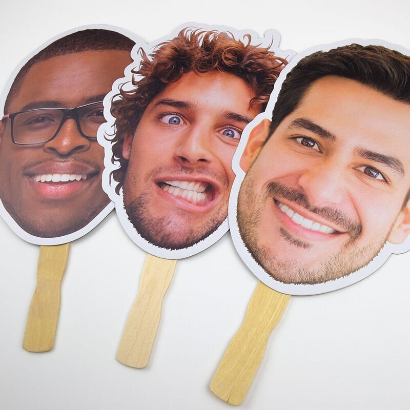 Photographs of three smiling men's faces on paper cutouts attached to wooden sticks.