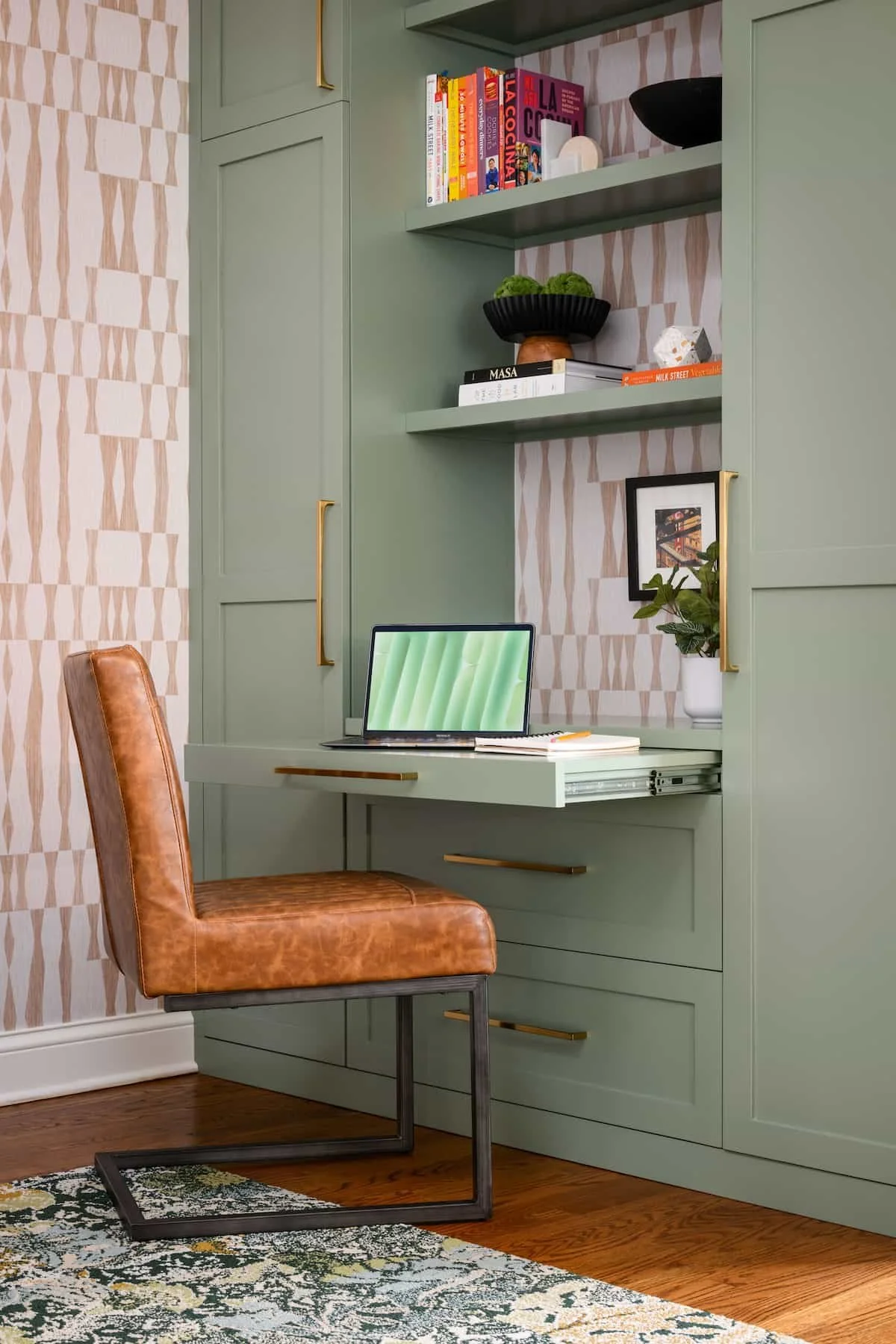 A home office corner featuring a built-in desk with a laptop, brown leather chair, and green cabinetry with gold handles. The shelves above hold books, decorative objects, and a framed picture. The wall has patterned wallpaper, and there is a patterned rug on the hardwood floor.