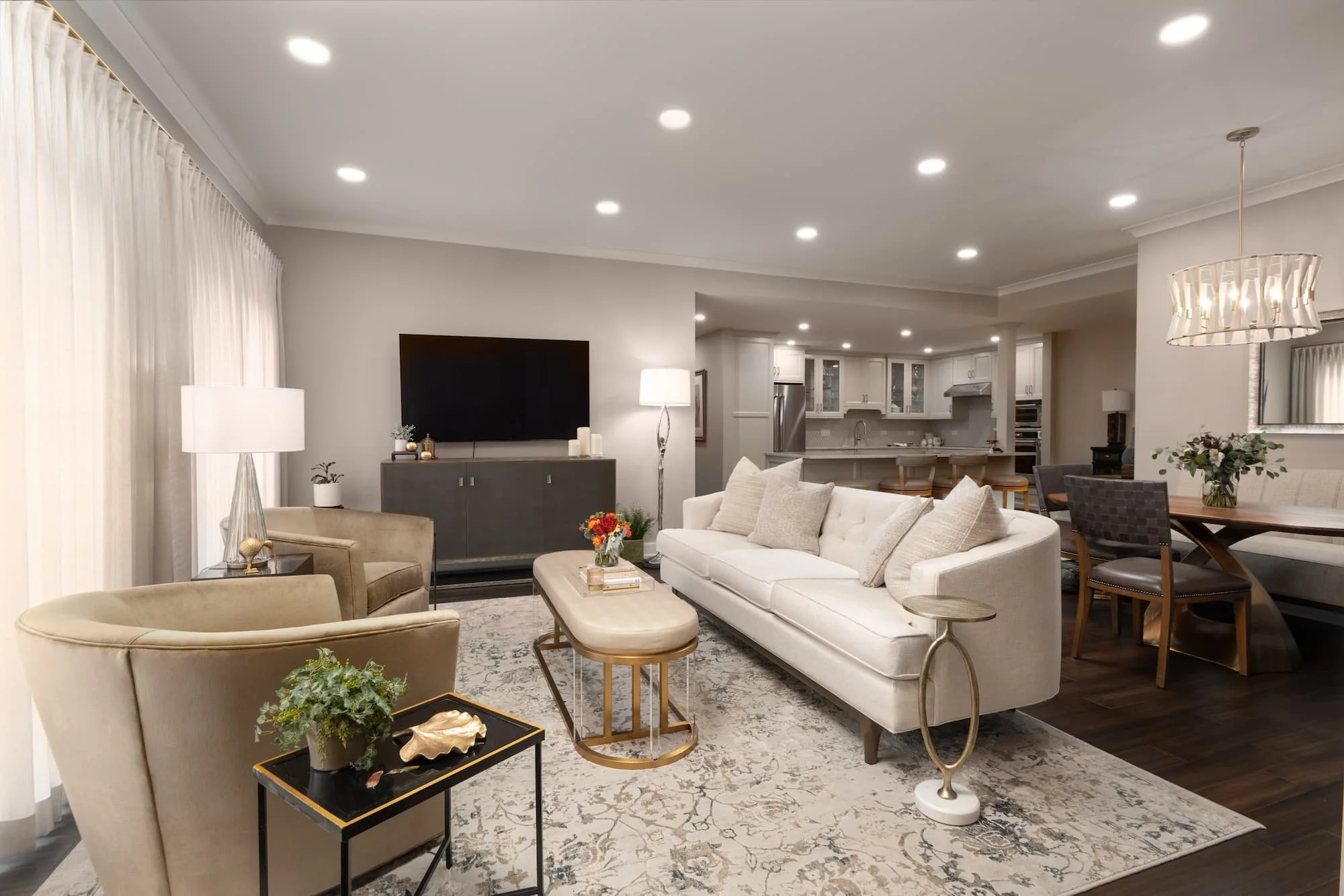 Spacious living room with cream-colored sofa, beige armchairs, dark wood coffee table, and a large patterned rug. Interior designed with wall-mounted TV, lamps, and decorative plants. Open plan layout leads to a modern white kitchen and dining area.