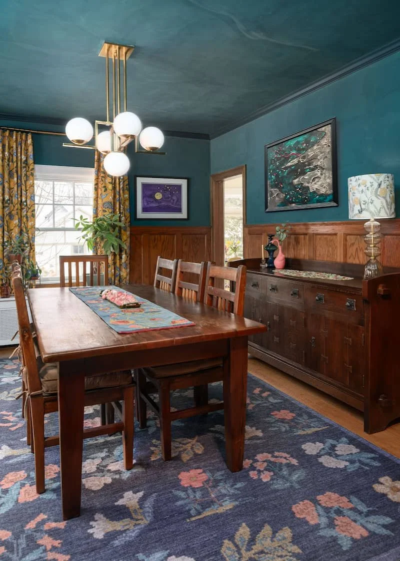 Dining room with midcentury modern globe lights above a craftsman style table. Yellow patterned curtains and emerald green walls accent a wood paneled room interior decorated in a colorful style.