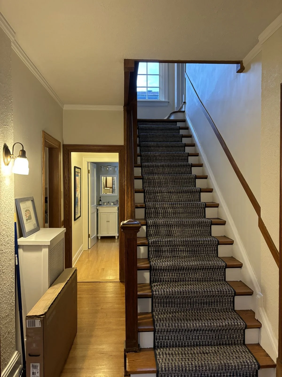 View of an interior staircase with a dark carpet runner, wooden steps, and a handrail, leading to a second story with a window at the top. To the left, an entryway with a white cabinet and a framed picture on top, a wall-mounted light fixture, and a doorway to another room. The floor is hardwood, and the walls are painted a light color.