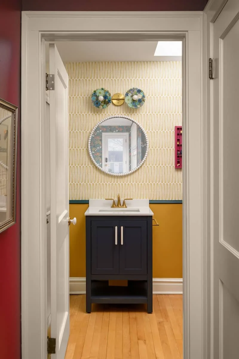 A small bathroom with yellow and white patterned wallpaper, a dark blue vanity with a white sink, a round mirror, a gold faucet, and decorative wall sconces with blue and white floral designs. There is a small red shelf on the wall to the right.