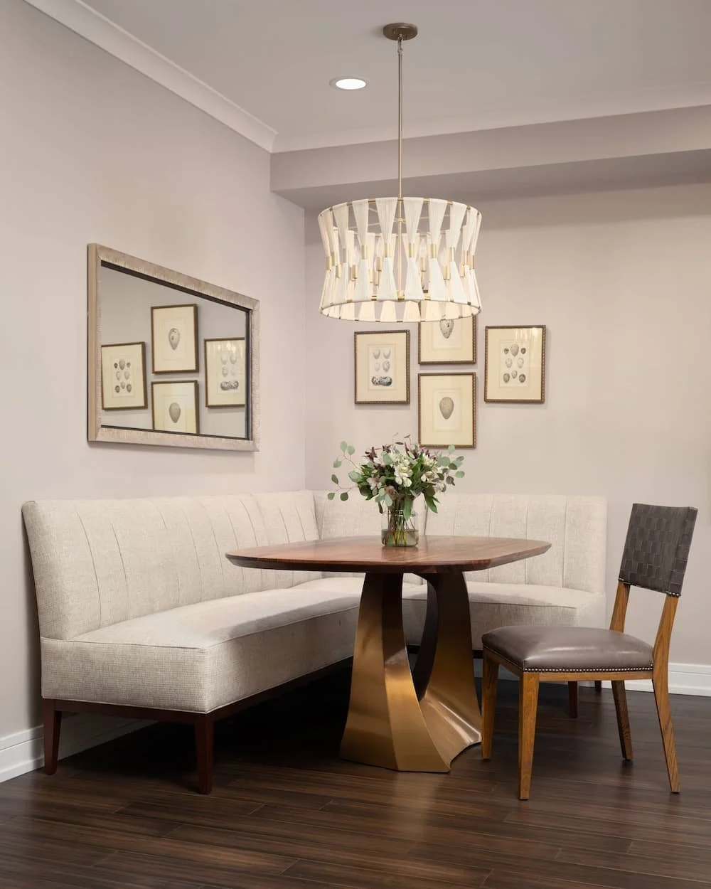 Dining nook with a beige upholstered corner bench, a wooden accent chair, a round wooden table with a gold base, and a floral centerpiece. A white chandelier hangs above, and framed artwork is on the wall.