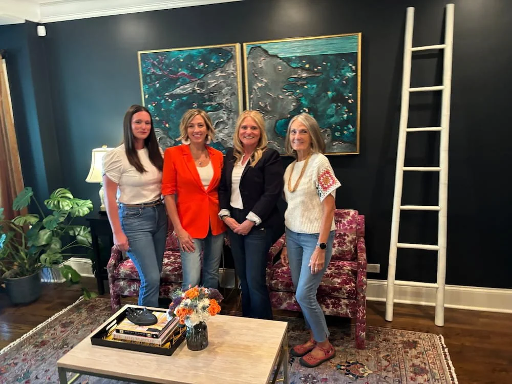 Four women standing together in a living room, smiling, with a dark wall, colorful artwork, a ladder, a floral couch, and a coffee table with a flower vase and books in the background.