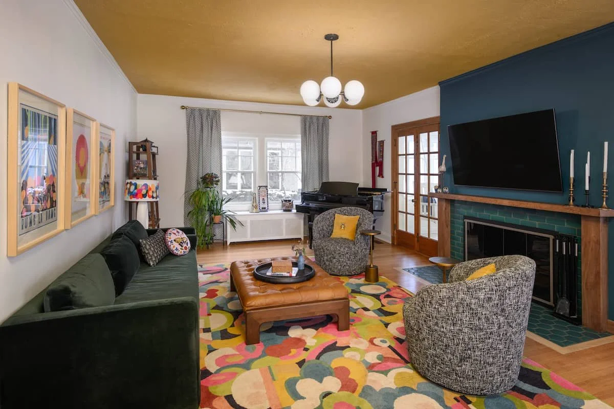 Living room with a green sofa, colorful patterned rug, two patterned armchairs with yellow pillows, a brown leather ottoman, a black piano near a window, a fireplace with a flat-screen TV above, and artwork on the walls.
