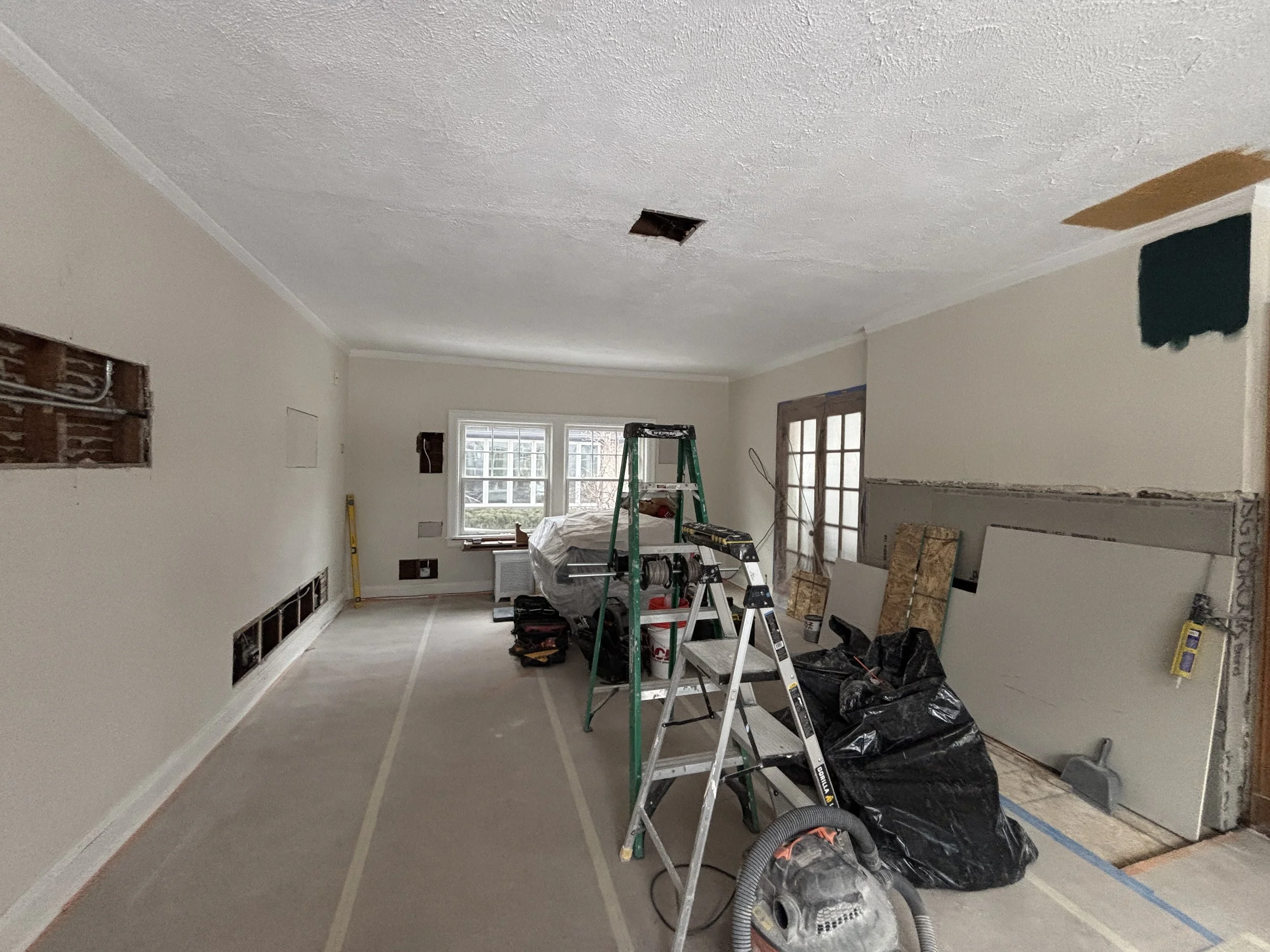 Living room under renovation with construction tools, ladders, and building materials scattered around. Part of the ceiling had a hole, and drywall is partially installed on one wall.