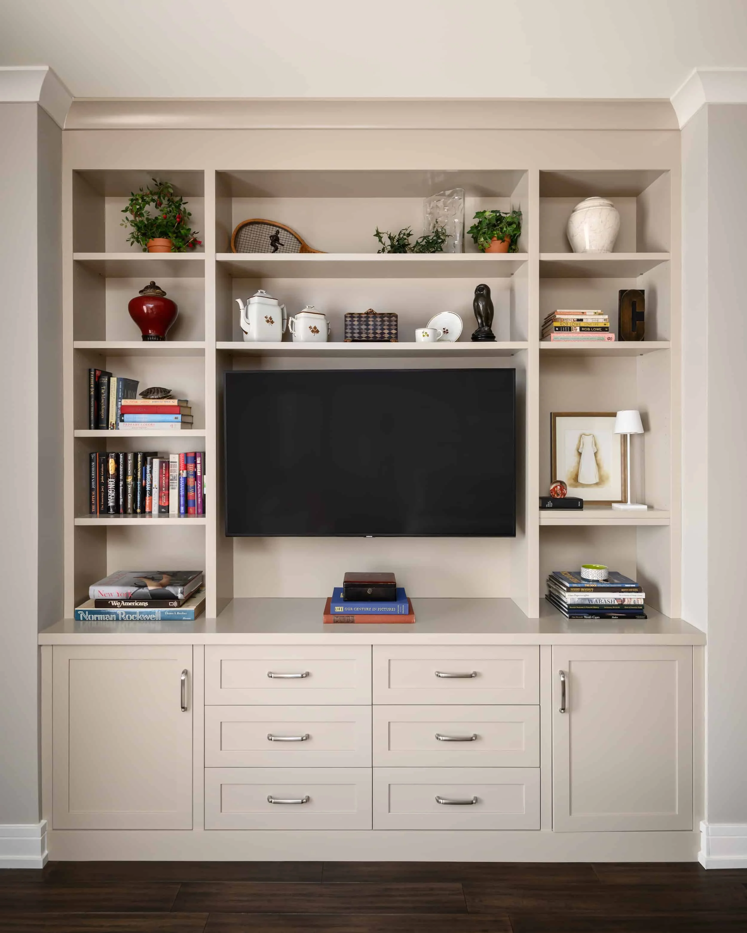Built-in white entertainment center with closed cabinets at the bottom and open shelves above, holding books, decorative items, and a large TV mounted in the center.