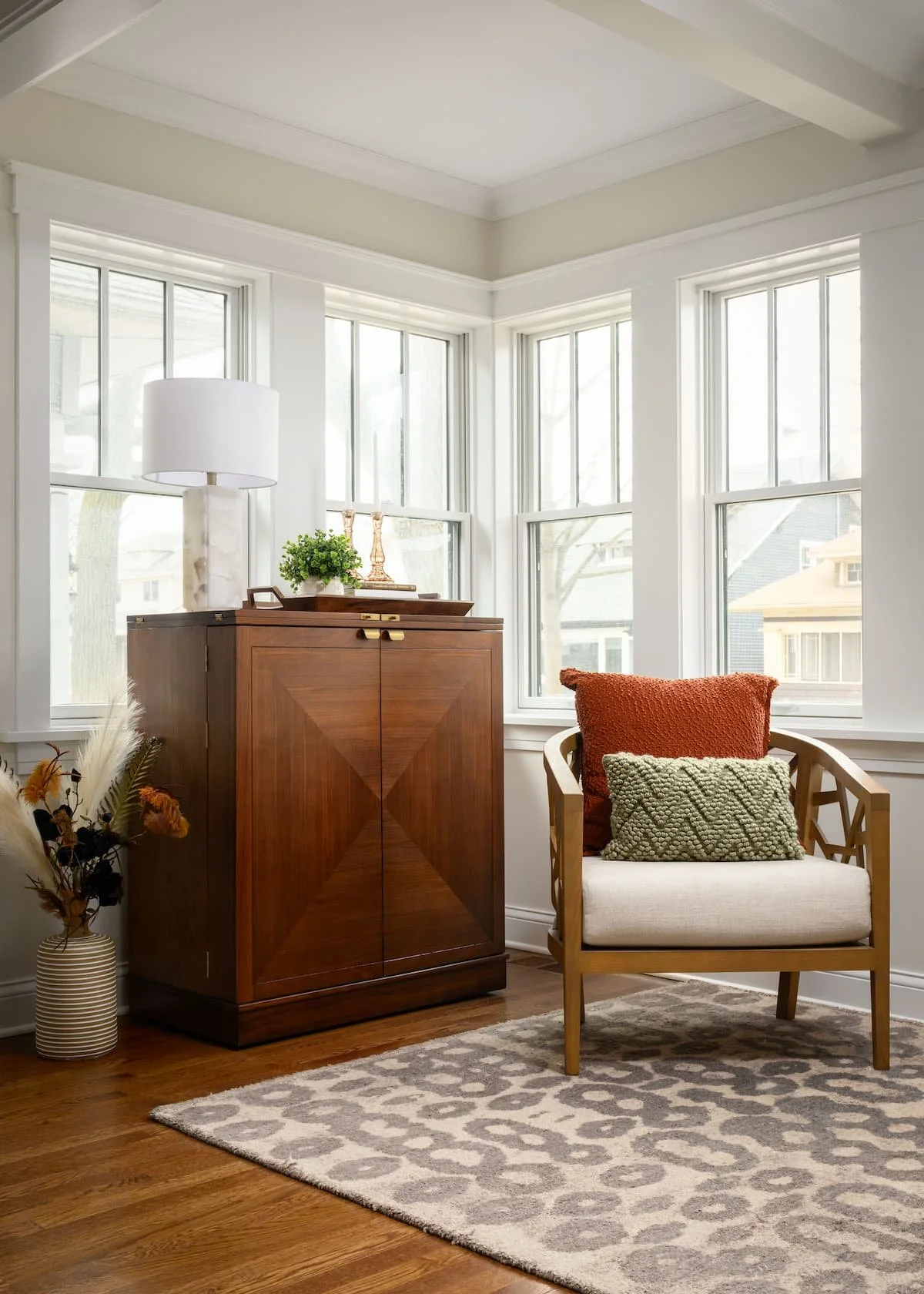 A cozy corner in a living room with a wooden armchair with red and green pillows, a wooden cabinet with decorative items on top, a lamp, and a vase with dried flowers, set on a wood floor with an area rug, in front of multiple large windows.