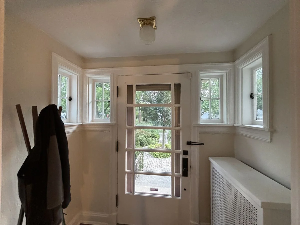 Entryway with front door and three windows, coat rack with jacket to the left, white walls, and a white radiator on the right.
