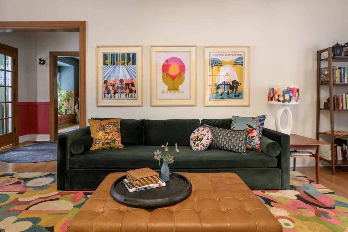 Living room with a dark green sofa, colorful pillows, framed artwork on the wall, a wooden side table with a patterned lamp, a bookshelf, and a brown leather ottoman with a tray and decorative items.