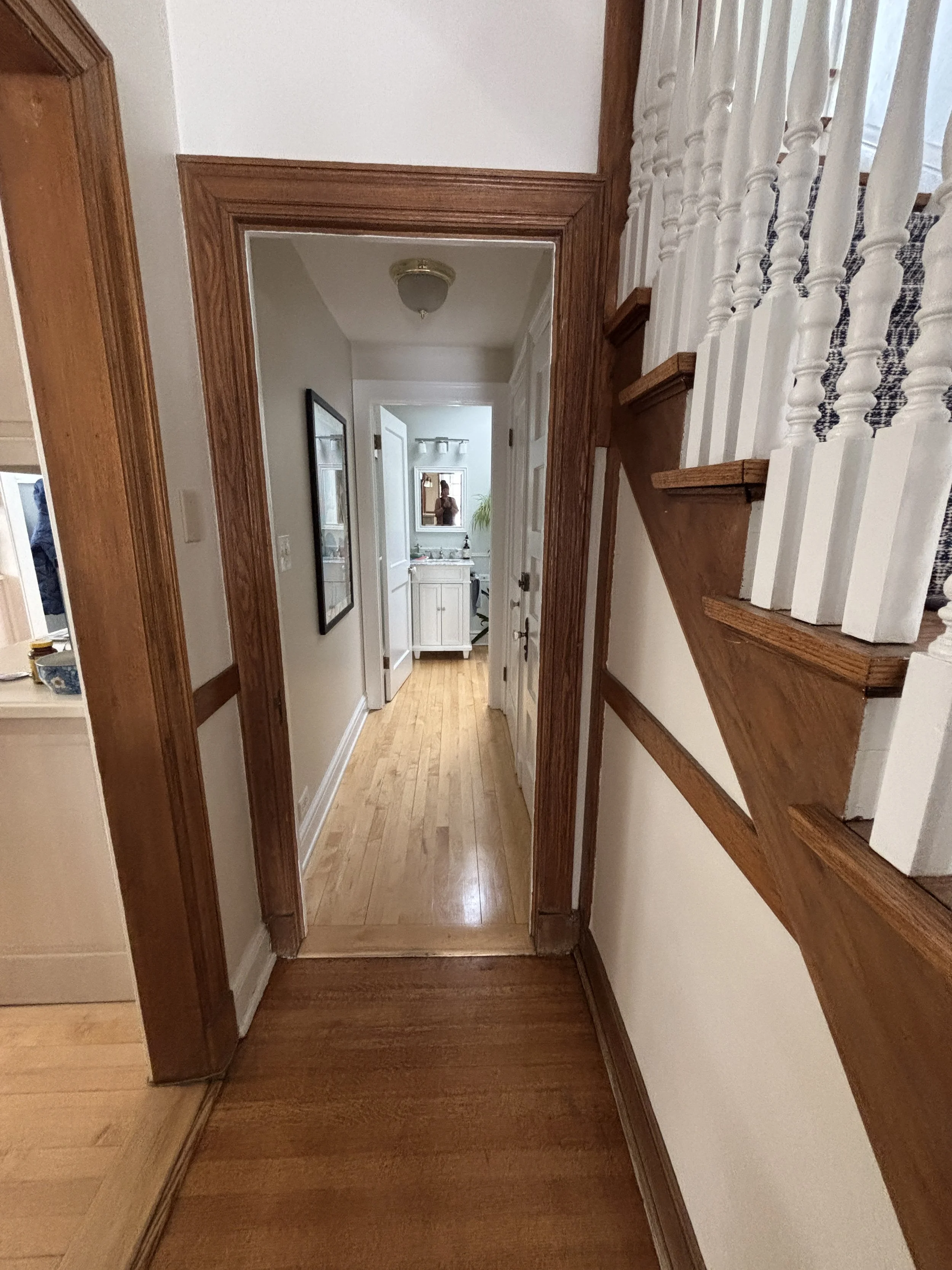 View down a hallway with hardwood floors, white walls, wood trim, and a staircase with white spindles and wooden steps on the right. At the end of the hallway, a bathroom with a white vanity, and a mirror.
