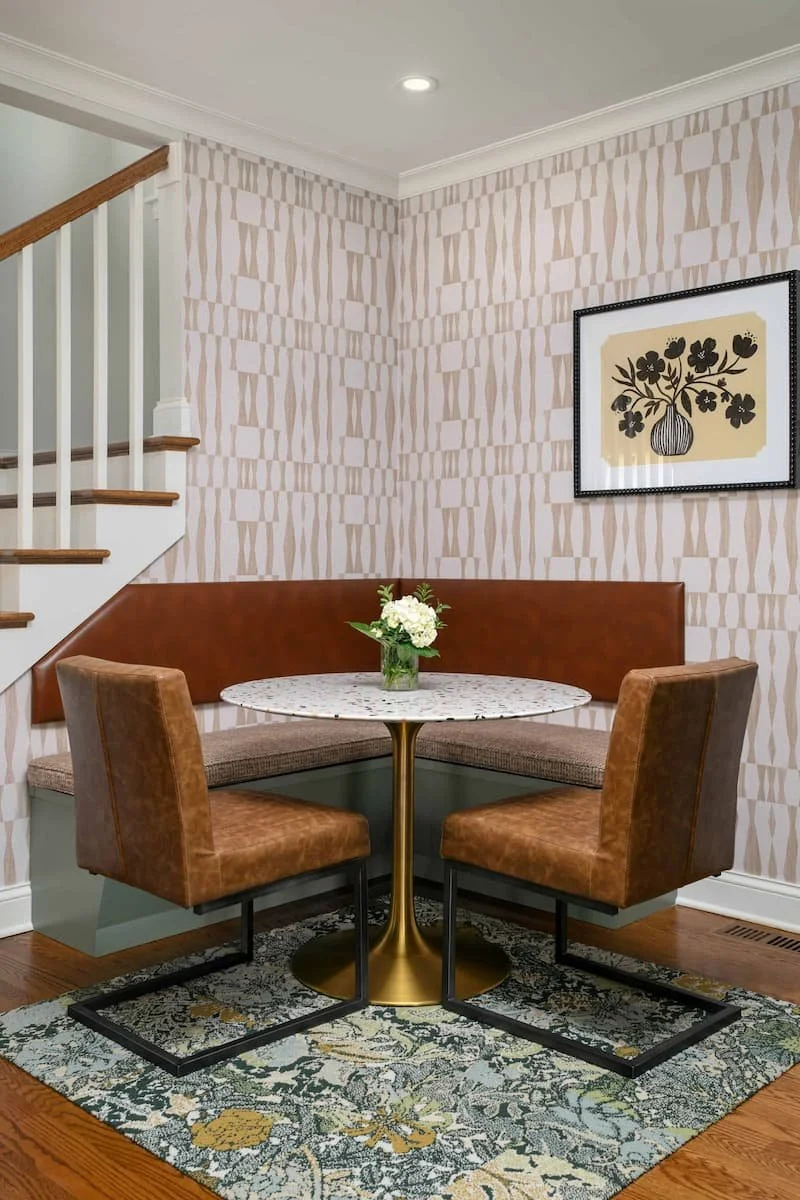 A cozy corner with a round table, lush floral centerpiece, upholstered chairs, patterned wallpaper, framed artwork, and a staircase in a home interior.
