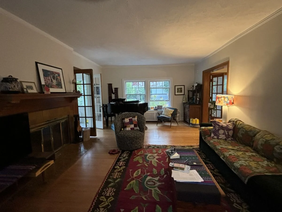 A cozy living room with a hardwood floor, a floral-patterned sofa, a patterned armchair, a colorful rug, a grand piano, a side table with a lamp, and large windows letting in natural light.