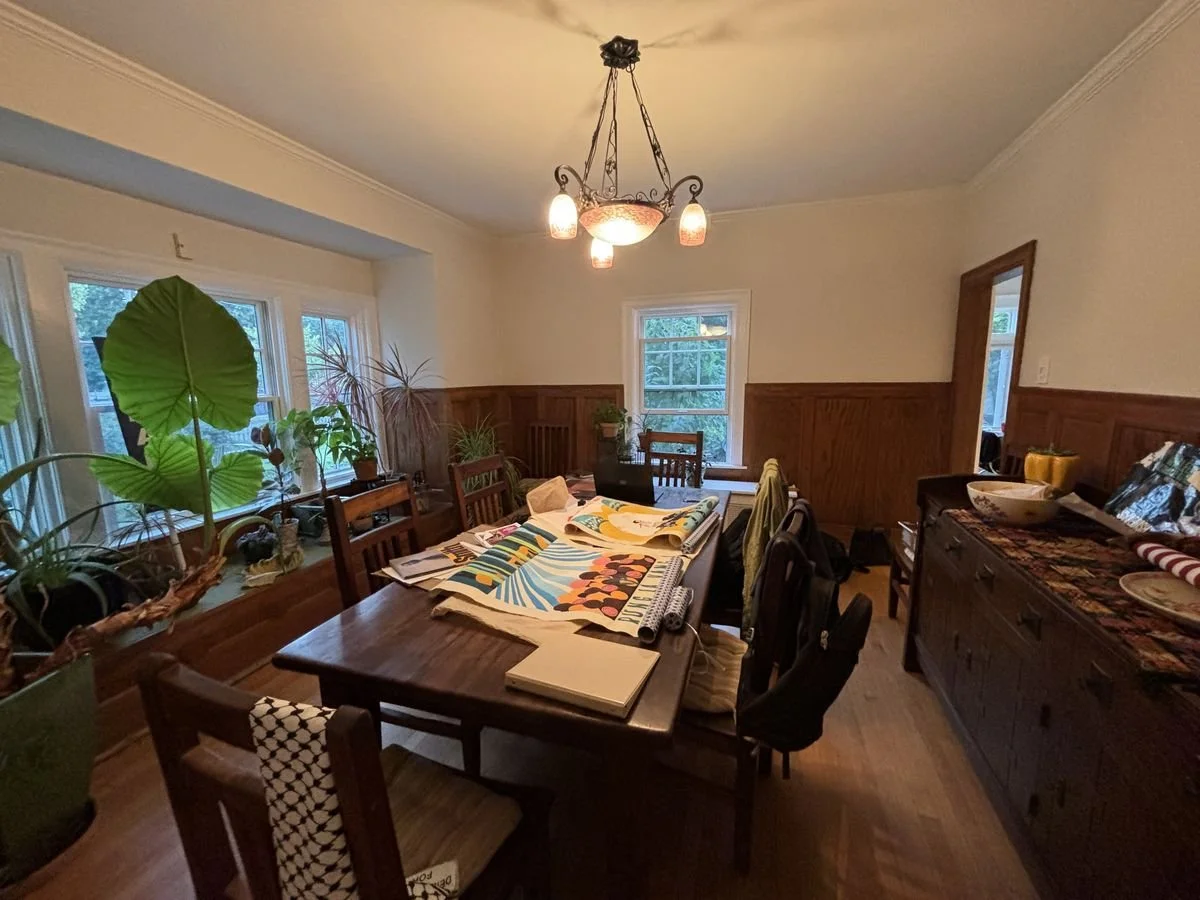 A dining room with a wooden table, chairs, and a sideboard. The table has art prints, a laptop, and notebooks. There are windows with plants and a ceiling chandelier.