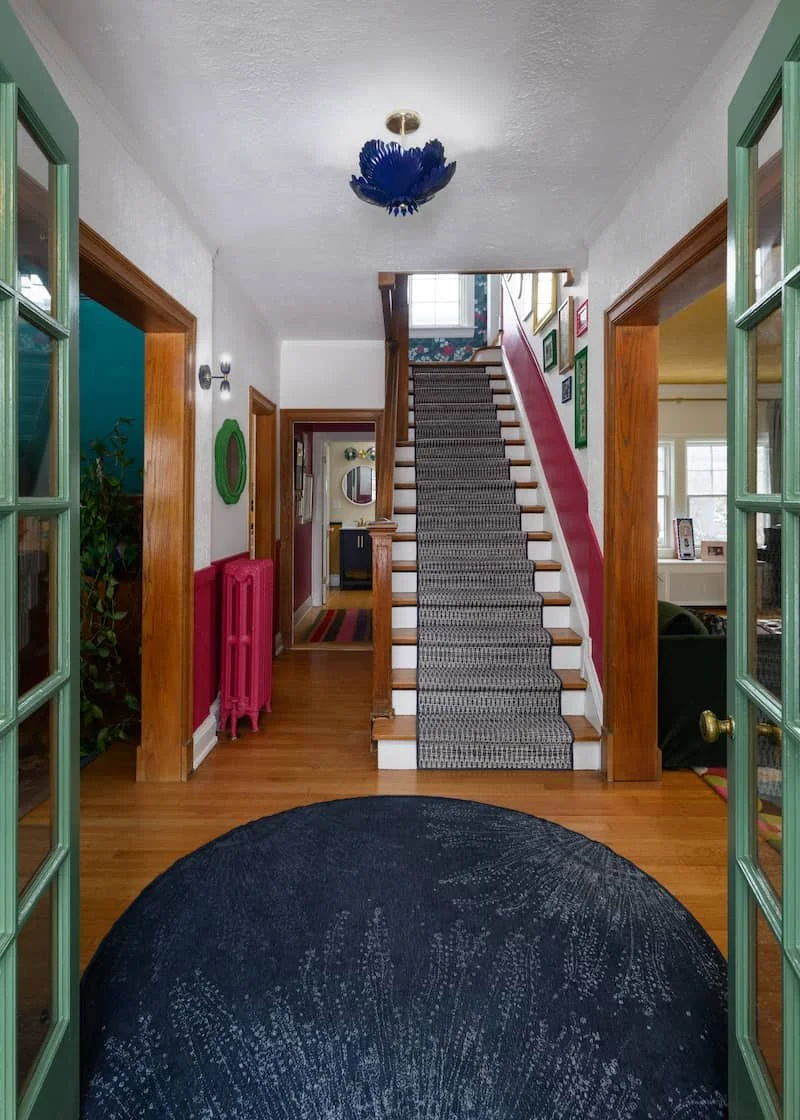 Interior view of a house entry hall with wood floors, a staircase with a patterned runner, and walls decorated with colorfully framed pictures. French doors with green trim open into the hall.