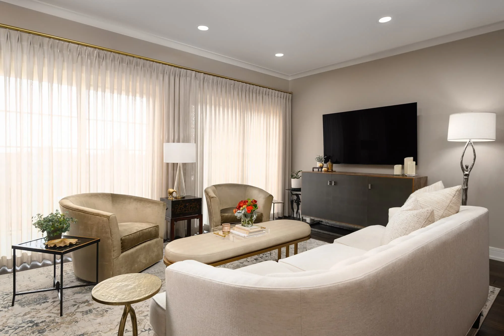 Living room with beige and cream furniture, a large flat-screen TV, curtains, lamps, and decorative plants.
