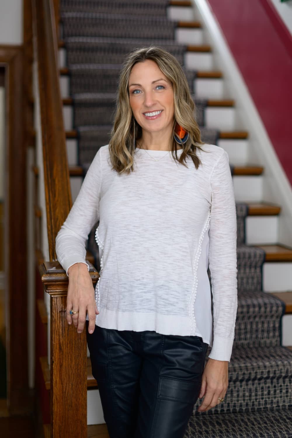 A woman with shoulder-length blonde hair, blue eyes, and wearing a white long-sleeve top and black pants, standing by a wooden staircase with a patterned carpet runner, smiling at the camera.