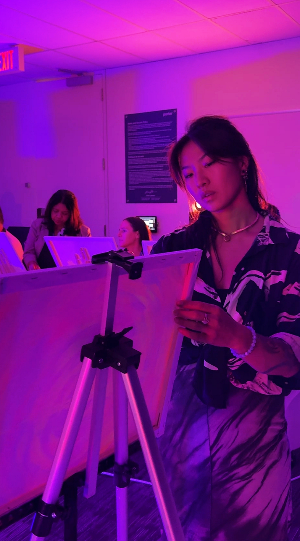 A woman in a patterned shirt standing in front of an easel, writing or drawing, in a room with purple and pink lighting, with other people in the background engaged in similar activities.