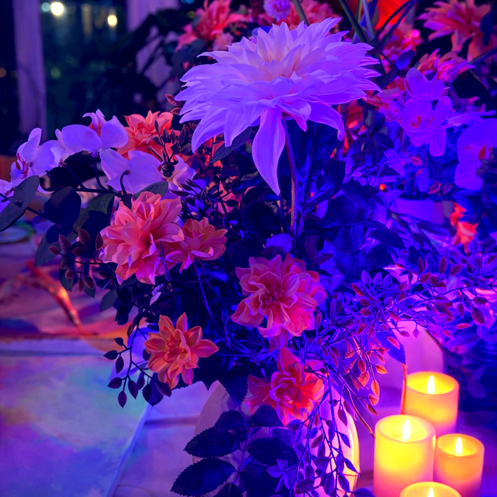 A colorful floral arrangement illuminated by purple and yellow lighting with candles in the foreground.