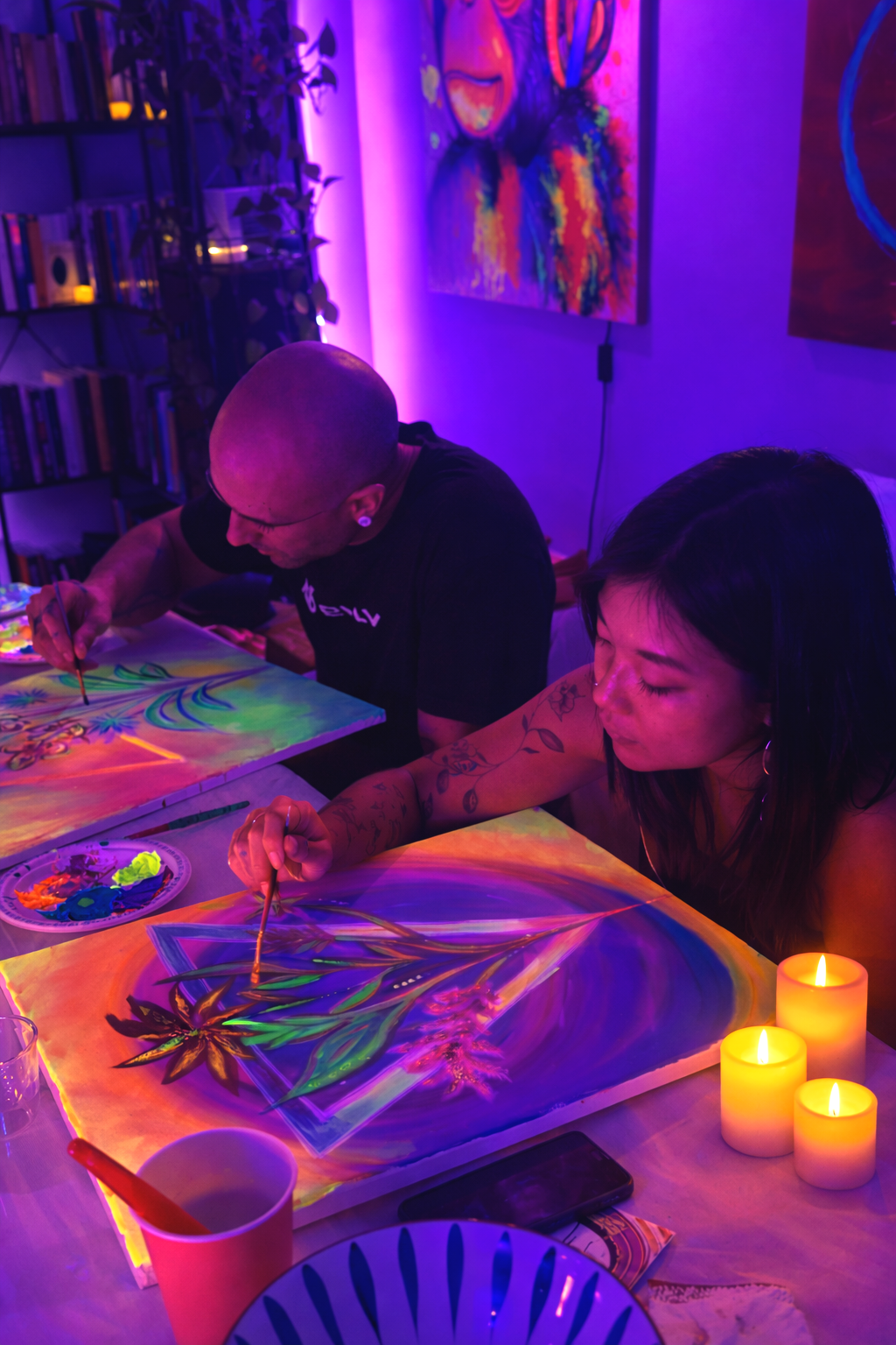 Two people creating neon glow-in-the-dark paintings under blacklight in a colorful, artistic space with candles and shelves of books in the background.