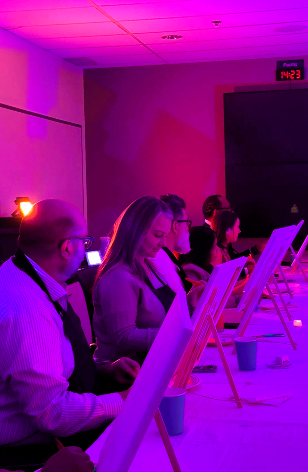 People participating in a painting or art activity in a dimly lit room with pink and purple lighting, each person focused on their easel.