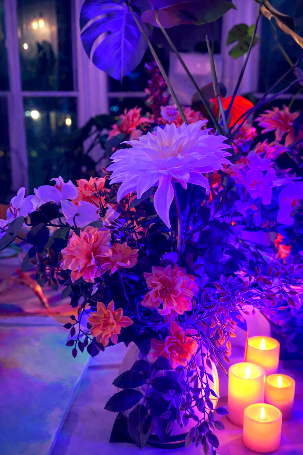 Colorful floral arrangement with large white and pink flowers illuminated by purple and yellow lighting, paired with four LED candles on a table.