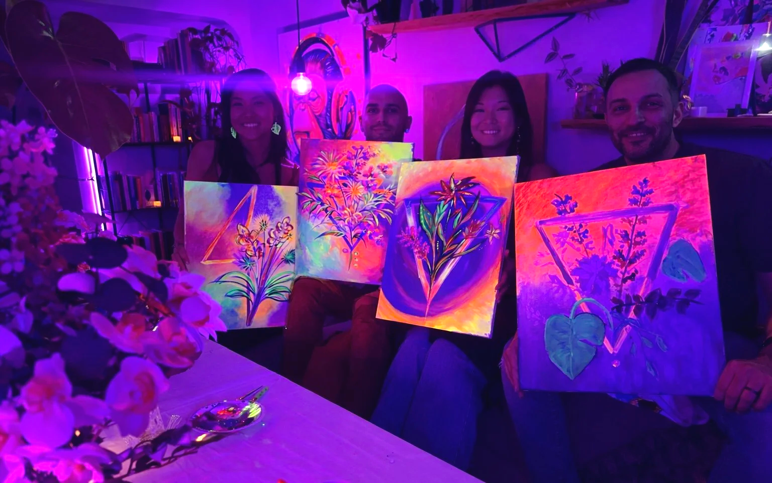 Four people sitting indoors, holding colorful glow-in-the-dark paintings of plants and flowers, in a room decorated with books, plants, and vibrant artwork.