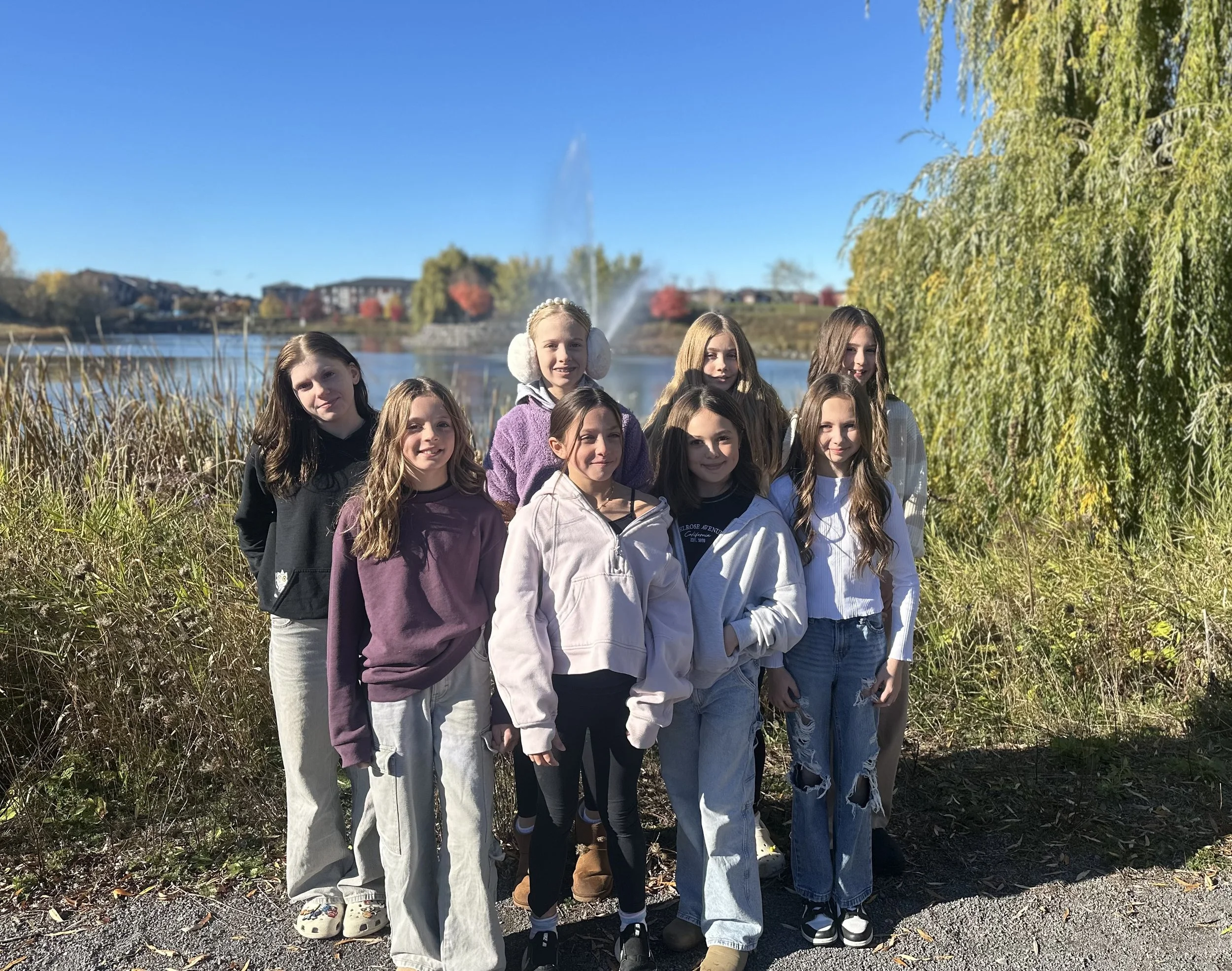 Group of 8 girls outside in various colored hoodies and pants. Background features trees and water with a fountain.